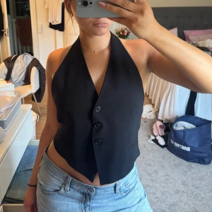 Svart västtopp med öppen rygg - Säljer en snygg svart västtopp med halterneck och knäppning framtill. Öppen i ryggen med ett band som går att anpassa hur tajt eller löst man vill. Från Zara Strl L men passar mig som har S/M.