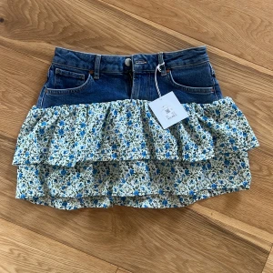 Jeanskjol med blommiga volanger - Supersöt kort jeanskjol med volanger i blommigt tyg. Kjolen har klassisk jeansmidja med fickor och knäppning, och volanger i vitt tyg med blå och gröna blommor. Perfekt för dig som vill sticka ut med en mix av denim och romantiska detaljer.