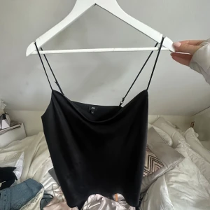 Svart satinlinne - Säljer en svart linnetopp från River island  i glansigt satinmaterial. Toppen har tunna, justerbara axelband och en snygg draperad urringning. Perfekt att styla med jeans eller kjol för en trendig look.