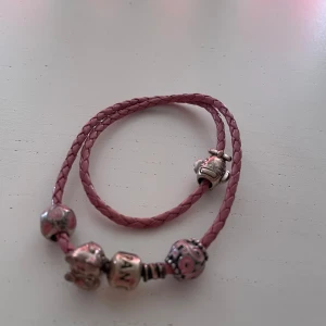 Pandora rosa läderarmband med berlocker - Dubbelrosettat armband i flätat rosa läder från Pandora, dekorerat med flera silverfärgade berlocker, bland annat en med hjärtan och en med mönster i rosa och silver. Perfekt för dig som gillar personliga smycken med charmig touch.