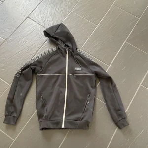 Svart hoodie från McKenzie - Svart hoodie från McKenzie med hel dragkedja framtill och vita detaljer. Jackan har huva med snörning, vita ränder över bröstet och längs ärmarna samt diskret mönster på axlarna. Två fickor med dragkedja och en bröstficka. Perfekt för en sportig och avslappnad stil.
