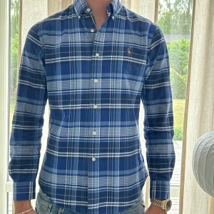 Blårutig skjorta från Ralph Lauren - Snygg blårutig skjorta från Ralph Lauren i slim fit-modell. Skjortan har klassisk krage, knäppning framtill och det ikoniska broderade logotypen på bröstet. Tillverkad i mjuk bomull och har långa ärmar. Perfekt för dig som gillar stilrena och tidlösa plagg.