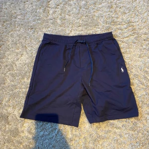 Marinblå shorts från Polo Ralph Lauren - Snygga marinblå shorts från Polo Ralph Lauren med klassisk broderad logga på benet. De har elastisk midja med snörning och är tillverkade i mjuk bomull, perfekta för sommardagar. Fickor på sidorna och en bakficka ger en clean och stilren look.               Om du skulle ha nån fråga så är det bara att skriva.