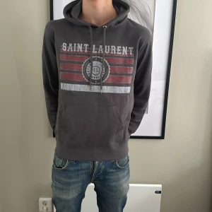 Saint laurent hoodie - Säljer en grå hoodie från Saint Laurent med vit och röd logga och tryck framtill. Tröjan har huva med snörning och en stor magficka. Skön passform och perfekt för dig som gillar streetstyle och designerplagg.