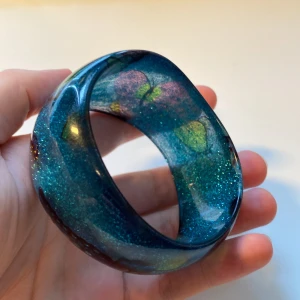 Glittrigt blått armband med fjärilar - Unikt armband i glittrig blå plast med inslag av lila och grönt. Armbandet har ett coolt fjärilsmotiv som syns tydligt och ger en färgstark vibe. Bred och rundad form som sticker ut och passar perfekt för dig som gillar statement-smycken.