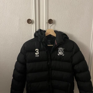 Svart pufferjacka från Vinson Polo Club - Svart pufferjacka från Vinson Polo Club med broderad logga och siffran 3 på bröstet. Jackan har huva, dragkedja och quiltad design. Perfekt för kalla dagar. 