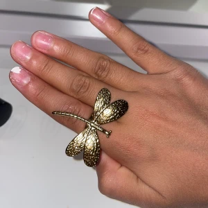 Guldig ring med trollslända - Cool statementring i guldmetall med en stor trollslända som motiv. Ringen har en bred, slät ringbas och trollsländan är detaljerad med prickig struktur på vingarna. Perfekt accessoar för dig som vill sticka ut och addera något unikt till din stil.