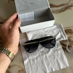 Svarta solglasögon från Dior - Snygga svarta solglasögon från Dior med markerad silvrig metallbåge upptill och mörka glas. Modellen har en modern, kantig form och logga på sidan. Kommer med originalbox, tygpåse och putsduk – perfekt för dig som vill sticka ut med stil.