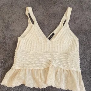 Vit stickad linnetopp med spets - Supersöt vit stickad linnetopp med v-ringning och breda axelband. Toppen har en volangkant i spets med broderade blommor längst ner som ger en romantisk känsla. Perfekt för varma dagar eller lager på lager.