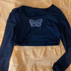 svart långärmad topp med fjäril i strass - Snygg svart croppad topp med långa ärmar och en glittrig fjäril framtill i strass. Toppen har en tight passform och är perfekt för dig som gillar edgy och trendiga detaljer. Materialet känns mjukt och stretchigt