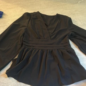 Svart omlottblus med v-ringning - Snygg svart omlottblus med v-ringad hals och långa ärmar med resår vid ärmslut. Blusen har markerad midja med breda band och ett lätt utsvängt fall. Perfekt för dig som gillar stilrena och trendiga plagg. Köpt second hand och det finns inget lapp men den sitter som en S. 