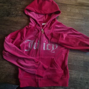  Juicy Couture kofta - Säljer en ikonisk zip-up  från Juicy Couture. Tröjan har huva, dragkedja med J-berlock och glittrande Juicy-text i strass på bröstet. Perfekt för dig som gillar 2000-talsvibbar och comfy stil.