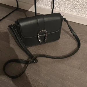 Helt Ny❤️ - Stilren svart axelväska i skinnimitation med justerbart axelband och snyggt silverfärgat spänne framtill. Väskan har en klassisk, rektangulär form och passar perfekt till en trendig vardagslook. Enkel design med fokus på detaljer.