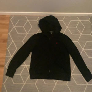 Svart hoodiejacka från Polo Ralph Lauren - Svart hoodiejacka från Polo Ralph Lauren med dragkedja framtill och klassisk huva. Jackan har en liten röd logga broderad på bröstet och är tillverkad i mjuk bomull. Perfekt för en chill och stilren look. Och är i strålande skick 