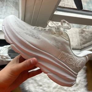 Vita Hoka träningsskor  - Snygga helvita sneakers från Hoka med luftig mesh-ovandel och riktigt tjock, dämpad sula. Skorna har snörning och diskreta logodetaljer på sidan. Perfekta för dig som gillar sportig stil och vill ha bekväma skor till vardags eller träning. Storlek 40 Finns i göteborg
