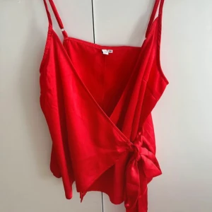 Röd omlottopp med knyt - Snygg röd omlottopp med smala axelband och knyt i sidan. Toppen har en v-ringad design och är tillverkad i ett mjukt, lätt tyg som ger en skön känsla. Perfekt för dig som vill sticka ut med en färgstark look