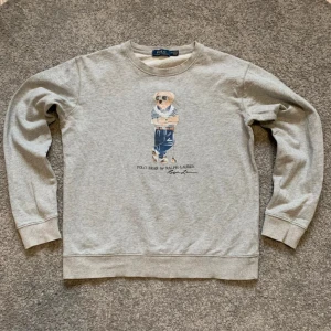 Grå sweatshirt med Polo Bear från Ralph Lauren - Säljer en grå sweatshirt från Ralph Lauren med det klassiska Polo Bear-motivet framtill. Tröjan har rund halsringning och långa ärmar. Perfekt för dig som gillar stilrena och ikoniska plagg. Storlek M men passar S