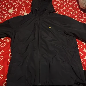 Svart vindjacka från Lyle & Scott L - Säljer en mörkblå vindjacka från Lyle & Scott i storlek L. Jackan har huva med dragsko, hel dragkedja framtill och den klassiska gula loggan på bröstet. Perfekt för blåsiga dagar och enkel att matcha med det mesta.