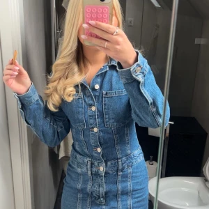 Kort jeansklänning med knappar - Snygg kort jeansklänning i blå denim med långa ärmar, knappar framtill och markerad midja. Klänningen har bröstfickor, krage och rå kant nertill för en cool vibe. Perfekt för dig som gillar en trendig och avslappnad stil.                    Första bilden är min, de andra två är från hon jag köpte den av!     Kan gå ner i pris, skicka förslag!