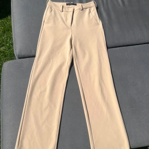 Beige kostymbyxor med raka ben - Snygga beige kostymbyxor med raka ben och klassisk passform. Byxorna har hällor för bälte, fickor i sidorna.Perfekta för en clean och stilren look.