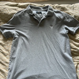 Grå pikétröja från Henri Lloyd - Snygg grå pikétröja från Henri Lloyd med klassisk krage och knappar framtill. Tröjan har korta ärmar och diskret broderad logga på bröstet. Perfekt för en clean och stilren look. Tillverkad i mjuk bomull för skön känsla hela dagen.