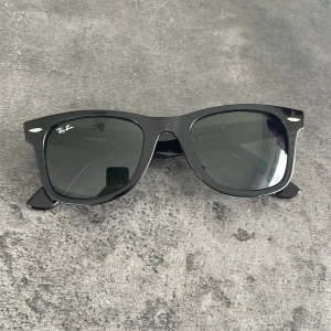 Svarta Ray-Ban Wayfarer solglasögon - Klassiska svarta Ray-Ban Wayfarer solglasögon med ikonisk fyrkantig form och mörka glas. Snygg och tidlös modell med Ray-Ban-logga på bågen. Perfekta för att lyfta din stil under soliga dagar.  Även äkta.