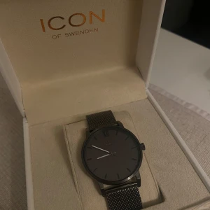 Svart klocka från Icon of Sweden - Stilren svart klocka från Icon of Sweden med mesh-armband i metall och minimalistisk urtavla utan siffror, förutom en romersk tolva. Klockan levereras i originalbox och har en modern, clean look som passar till många outfits.