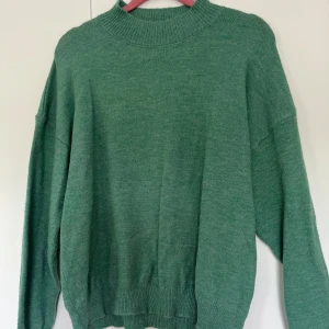 Grön stickad tröja med halvpolo - Mysig grön stickad tröja med hög ribbad krage, nästan halvpolo och långa ärmar. Tröjan har en loose passform och breda muddar vid ärmslut och nederkant. 
