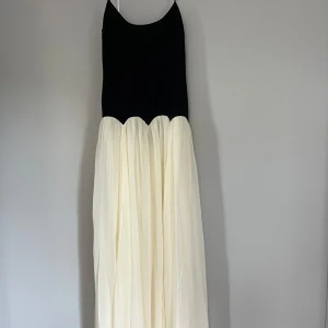 Svart och vit långklänning med öppen rygg - Supersnygg långklänning med svart figurnära överdel och tunn vit kjol. Klänningen har smala axelband, öppen rygg med snörning och en vågig övergång mellan överdel och kjol. Perfekt för dig som vill sticka ut med en elegant och modern look.