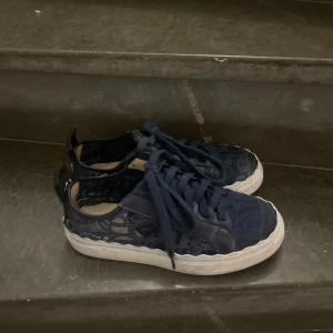 Mörkblå sneakers från Chloé i spets - Snygga mörkblå sneakers från Chloé med cool spetsdetalj och vit platt sula. Skorna har snörning och tydlig Chloé-logga baktill. Ovandelen är i textil med läckert mönster och läderdetaljer. Perfekta för dig som vill sticka ut med en unik och trendig look. Bra skick. 