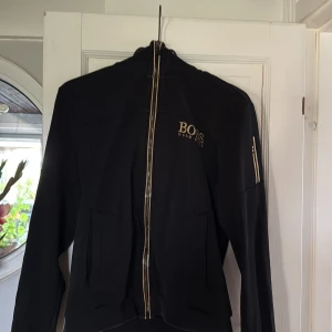 Svart vindjacka från Hugo Boss - Svart vindjacka från Hugo Boss med guldig logga på bröstet och dragkedja framtill. Jackan har hög krage, långa ärmar och diskreta fickor. Snygga guldiga detaljer på ärmen och dragkedjan ger en exklusiv känsla. Perfekt för dig som gillar stilrena och sportiga plagg.