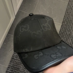 Svart keps från Gucci med GG-mönster - Snygg svart keps från Gucci med klassiskt GG-mönster över hela kepsen. Kepsen har en böjd skärm och är tillverkad i ett slitstarkt syntetmaterial med diskret glans. Perfekt accessoar för att lyfta din streetwear-look. Kommer med boxen. Och är helt ny har bara haft den i en vecka.