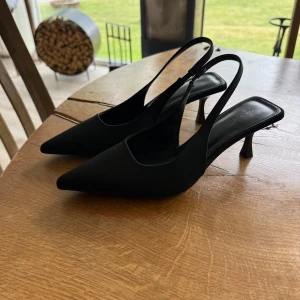 Svarta pumps med spetsig tå från nakd  - Stilrena svarta pumps från nakd med spetsig tå och smal klack. Skorna har öppen häl med smal rem runt ankeln och är tillverkade i ett slätt material som ger en elegant look. Perfekta för dig som gillar minimalistisk och trendig stil.