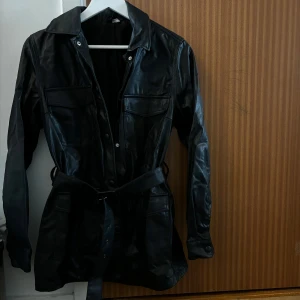 Svart skinnjacka med bälte - Snygg svart skinnjacka med bälte i midjan och stora fickor framtill. Jackan har klassisk krage, knappar och långa ärmar. Perfekt för dig som vill ha en edgy och stilren look.