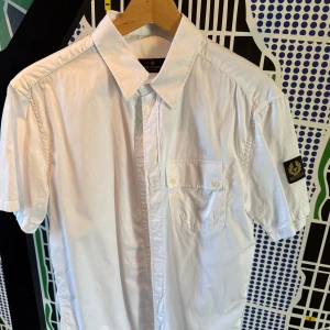 Vit kortärmad skjorta från Belstaff - Vit skjorta från Belstaff i 100% bomull med korta ärmar och klassisk krage. Skjortan har en bröstficka med knapp och en snygg Belstaff-logga på ärmen. Perfekt för dig som gillar stilren och enkel design.