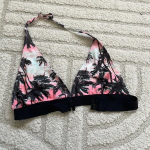 Palmmönstrad bikiniöverdel i rosa - Snygg bikiniöverdel med palmmotiv i svart mot en bakgrund i rosa, vitt och ljusblått. Triangelformade kupor och svart band under bysten med spänne bak. Perfekt för stranden eller poolen.