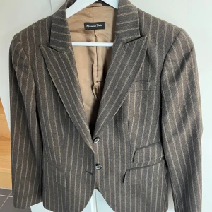 Randig brun kavaj från Massimo Dutti - Snygg brun kavaj från Massimo Dutti med klassiska smala ljusa ränder. Kavajen har två knappar framtill, fickdetaljer och är helfodrad i beige. Perfekt för dig som gillar en tidlös och stilren look.
