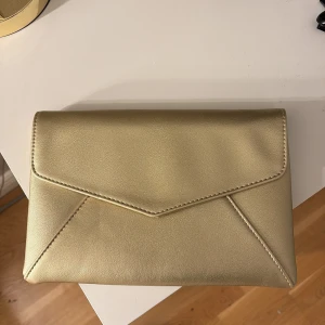 Guldig kuvertväska från Anna Field - Snygg kuvertväska i guld från Anna Field med stilren design och magnetlås. Väskan är gjord i syntetmaterial och har ett innerfack för småsaker. Aldrig använd☺️