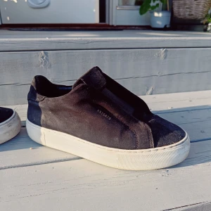 Svarta sneakers från Axel Arigato - Stilrena svarta sneakers från Axel Arigato med minimalistisk design och vit platt sula. Ovandelen är i mocka och skon har en clean look utan synlig snörning. Perfekta för dig som gillar en modern och enkel stil.