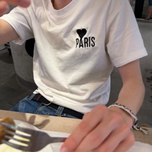 Ami paris tröja - Vit t-shirt i bomull med svart hjärta och texten 'PARIS' tryckt på bröstet. Klassisk rund halsringning och korta ärmar. Enkel och stilren design som passar perfekt till jeans eller shorts.