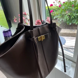 Brun axelväska i skinn med gulddetalj - Snygg brun axelväska i skinn med stilren design och guldfärgat spänne framtill. Väskan har långa handtag och rymligt innerfack med dragkedja. Perfekt för dig som vill ha plats för allt du behöver under dagen.