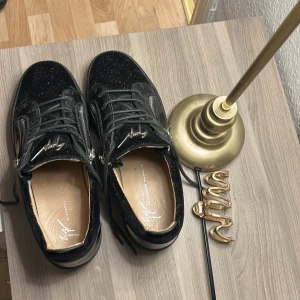 Svarta sneakers från Giuseppe Zanotti - Snygga svarta sneakers från Giuseppe Zanotti med glittrig finish och coola silverdetaljer. Skorna har snörning, dragkedjor på sidorna och en platt sula. Materialet är en mix av mocka och syntet, med exklusiv känsla och ikonisk logga på plösen.