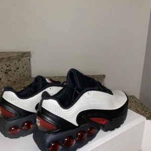 Nike DN Shox vita och svarta sneakers  - Nike DN Shox sneakers i vitt och svart med röda detaljer och ikoniska fjädrande Shox-kulor i sulan. Ovandel i syntetmaterial med mesh för andningsförmåga, svart snörning och rund tå. Perfekta för dig som vill sticka ut med en futuristisk vibe.
