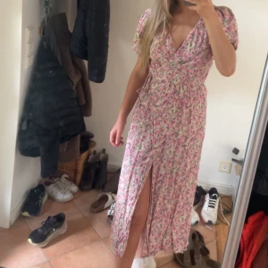 Blommig omlottklänning med slits - Söt långklänning med blommigt mönster i rosa, vit och grönt. Omlottmodell med v-ringning, korta puffärmar och en snygg slits framtill. Perfekt för sommarens alla tillfällen och ger en romantisk vibe.