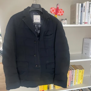 Svart Moncler dunblazer - Snygg svart dunjacka från Moncler i blazer-modell med tre knappar framtill, klassisk krage och flera fickor. Jackan har quiltad insida och en stilren look som passar perfekt när du vill vara både varm och uppklädd. Fodret är glansigt och jackan har lång ärm.