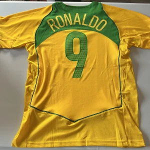 Brasilien Ronaldo 9 Nike fotbollströja - Gul Brasilien fotbollströja med gröna detaljer, nummer 9 och Ronaldo på ryggen. Klassisk Nike-design med broderat landslagsemblem och gröna inslag vid krage och sidor. Tillverkad i lätt och ventilerande polyester, perfekt för match eller träning. Replika. Endast handtvätt gäller!