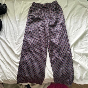 Mönstrade vida byxor från Zara - Färgstarka vida byxor från Zara med ett coolt geometriskt mönster i lila, blått och orange. Byxorna har resår i midjan och är tillverkade i ett glansigt material som känns lyxigt. Perfekta för dig som vill sticka ut med din stil.