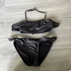 Svart bikini med volang från H&M - Svart bikini från H&M med volangdetalj på överdelen och knyt i sidorna på underdelen. Axelbandet har en liten guldfärgad snäcka som extra detalj. Snygg och stilren modell, perfekt för stranden eller poolen.