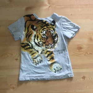 Grå t-shirt med tigertryck H&M x Nathalie Lété - Grå t-shirt från H&M x Nathalie Lété med stort, färgstarkt tigertryck och texten 'Le Fabuleux Tigre' på framsidan. Klassisk passform med korta ärmar och rund hals. Mjuk bomullskvalitet, perfekt för dig som gillar djurmotiv och coola prints.