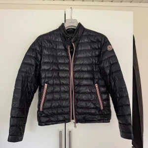 Svart Moncler dunjacka - Snygg svart dunjacka från Moncler med klassiska röda och vita detaljer på dragkedjorna. Jackan har hög krage, två fickor med dragkedja och Moncler-logga på ärmen. Perfekt för höst/vår. Storlek 2. Fint skick. Pris kan absolut diskuteras. 